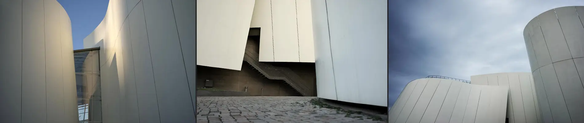 Hochwertige Fotografie moderner Architektur: Geschwungene, glatte Oberflächen, geprägt von klaren Linien und organischen Formen. Eine schmale Treppe führt zwischen den Wänden empor, während das historische Kopfsteinpflaster einen spannenden Kontrast schafft. Die präzise Bildkomposition unterstreicht die Eleganz und den minimalistischen Charme des Bauwerks.